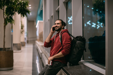 Fototapeta premium A man is talking on the phone at the airport at night. A guy, a tourist with a hand luggage, is waiting for departure at the gate