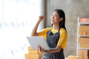 Naklejka premium Small business owner in Asian woman who are delighted with successful looking at laptop. Excited Success. Asian businesswoman raising hand.