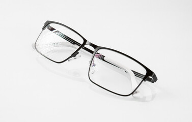 Glasses in a black metal frame are reflected in a white mirror background and have glare on the lenses