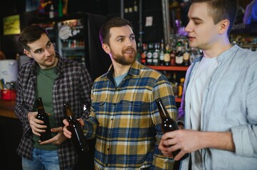 Three young men in casual clothes are smiling, holding bottles of beer while standing near bar counter in pub