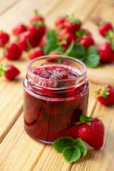 Strawberry confiture in glass jar on wooden board with fresh strawberry fruit and green leaves on wooden background. Recipe of homemade berry jam of strawberry full of vitamins and antioxidants.