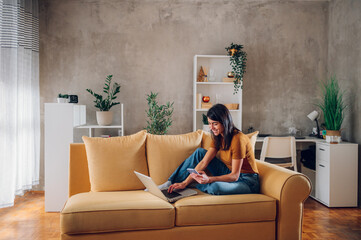 Woman using laptop and a smartphone while sitting on a yellow sofa at home
