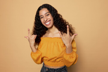 Cheerful latin dark-skinned girl in summer clothes showing rock star gesture with hands, looking at camera with toothy smile, feeling cool and happy. Human emotions and feelings, body language