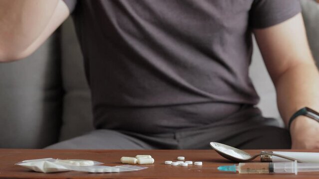 Addicted man sitting on a sofa, drinking alcohol from a glass and taking drugs and pills on a table with syringe and white powder narcotics