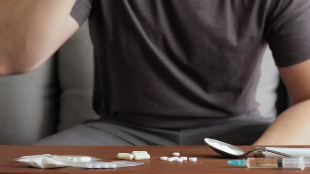 Addicted man sitting on a sofa, drinking alcohol from a glass and taking drugs and pills on a table with syringe and white powder narcotics