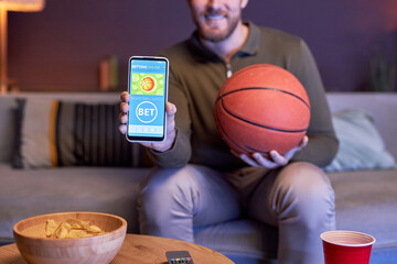 Close up of man holding smartphone with online sports betting on screen and basketball ball, copy space