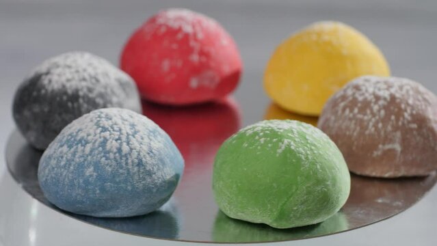Six brown, green, red, black, yellow, and black mochi cakes spin while lying on a mirror surface. Presentation of cake flavors on a light background.