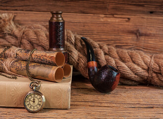 Stare, eleganckie przedmioty, zwoje map i zegarek kieszonkowy leżące na książce oraz luneta, fajka do tytoniu i gruba lina na drewnianym tle. 