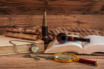 Vintage items, old books, old maps, tobacco pipe, magnifying glass, telescope, rope, old keys and pocket watch lying on wooden boards.