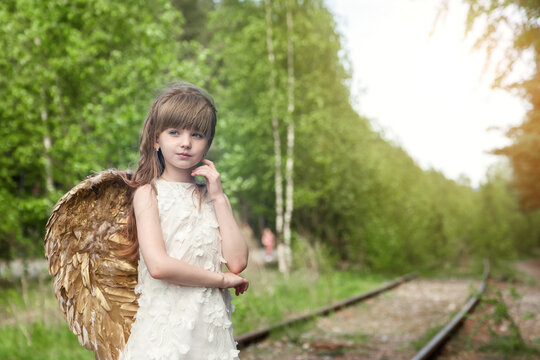 Pensive little girl in angel image with gold wings posing in greenery forest, looking away. Thoughtful young lady in white dress in sunshine woodland. Fairytale, fantasy concept. Copy ad text space