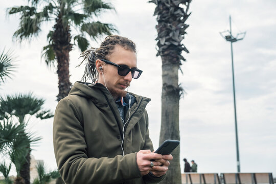 Young Caucasian man with dreadlocks listening to music with earphones using smartphone outdoor