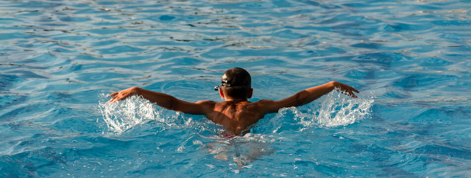 Back view of young athlete swim in outdoor swimming pool on summer sunny day. Boy child swimmer swim butterfly style. Wide banner