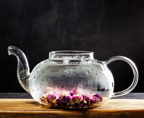 A beautiful glass teapot for brewing tea stands on a wooden board on a dark background. The teapot...