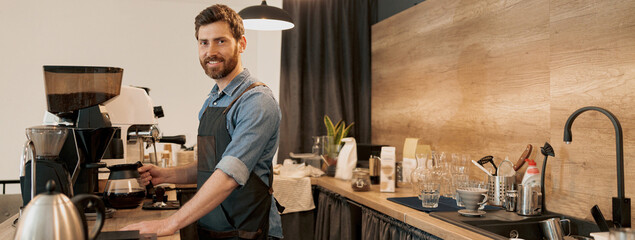 Smiling barista with stylish beard holding glass with filter coffee standing behind the counter