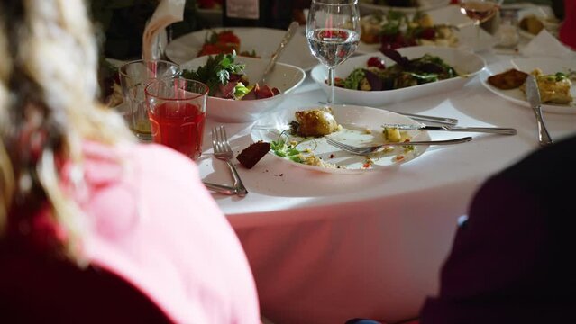 Empty Dirty Plate After Meal On Served Banquet Table With Wine Glasses With Water And Salads. Older Couple Is Sitting At Table, Sunlight Falls On Table And On Woman.