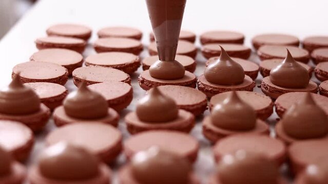 Making french desert macarons. Chef hands with confectionery bag add cream to the ready halves of brown chocolate macaroons at pastry shop. cooking, food and baking concept