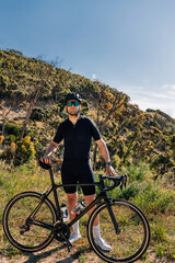 Fototapeta premium Male cyclist with bicycle in wild terrain looking at camera. Athlete in black sportswear with road bike relaxing.