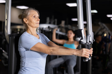 Fototapeta na wymiar Concentrated motivated sporty elderly woman exercising chest muscles on butterfly machine during upper body strength training in gym