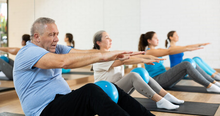 Portrait of active senior man during group core training, sitting on mat and doing sit-ups with bender ball at gym. Healthy lifestyle and pilates concept