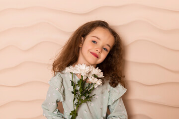 Portrait of a charming little girl, well dressed in a pastel green dress with a slight makeup and brown loose hair posing with a flowers on a short bench against a wavy beige wall.