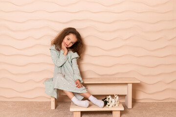 Portrait of a charming little girl, well dressed in a pastel green dress with a slight makeup and brown loose hair posing with a flowers on a short bench against a wavy beige wall.