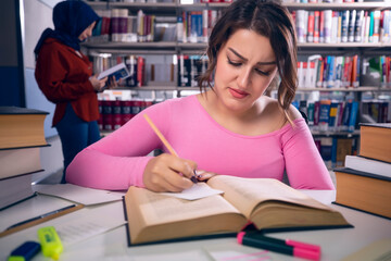 Young, beautiful student girl who studies at the library and writes notes. The girl is having a hard time studying.