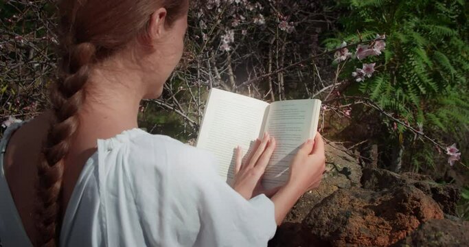 Woman reads book outdoor. Rear view of girl student studying information in spring blooming garden.