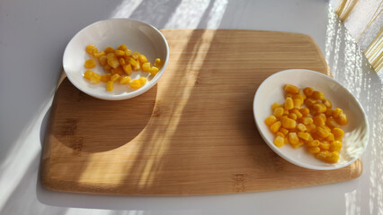 Lot of pieces of canned yellow corn on plate which is on wooden bamboo cutting board on white background. The concept of cooking and delicious healthy food