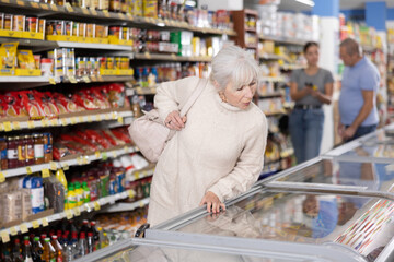 Attentive old woman purchaser choosing frozen product out of large stock in supermarket