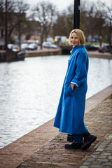 A woman in a long blue coat in the Netherlands. Tourist while traveling in Rotterdam. An adult woman near the canal.