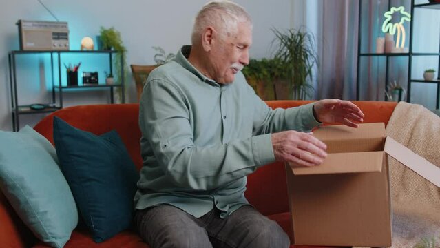 Happy senior grandfather man unpacking delivery parcel sits at home. Smiling satisfied elderly guy shopper, online shop customer opening cardboard box receiving purchase gift by fast postal shipping