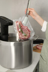 Woman putting vacuum packed meat into pot with sous vide cooker in kitchen, closeup. Thermal...