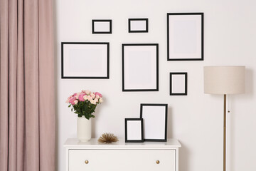 Fototapeta premium Empty frames hanging on white wall and chest of drawers with flowers indoors
