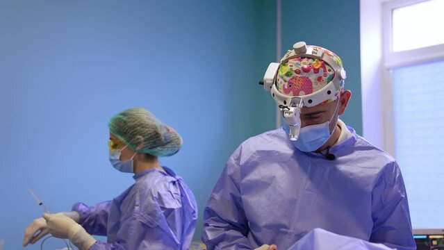 Calm serious surgeon performs operation in surgery room. Nurse prepares tools at backdrop.