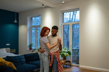 Joyful young diverse married couple dancing in living room at home, happy multiracial family man...