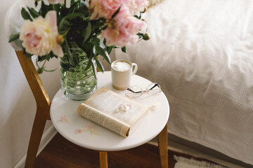 Still life details in home interior of living room. Open book with glasses, cup coffee and bouquet white pink peonies flowers. Read and rest. Cozy home
