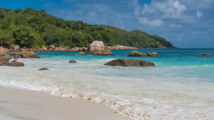 Fototapeta na wymiar The waves of the turquoise ocean foam on the white sand of the beach. Picturesque boulders in the surf. A hill overgrown with tropical vegetation against a blue sky and clouds. Seychelles. Praslin.
