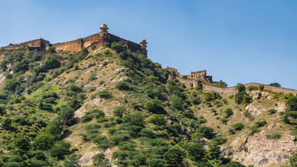 Fototapeta na wymiar The ancient fortress wall runs along the ridge of the mountain. Watchtowers with domes are visible. Blue sky. Green vegetation on the slope. India. Amber Fort. Jaipur