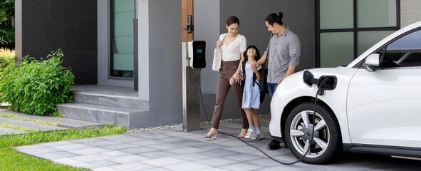 Progressive young parents and daughter with electric vehicle and home charging station. Green and...