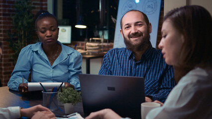 Employees coworking at boardroom table in business meeting, woman holding report clipboard, man working on laptop, looking at camera. Diverse colleagues team brainstorming