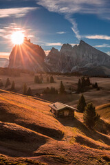 Alpe di Siusi, Italy - Autumn sunrise with a wooden chalet at Seiser Alm in South Tyrol in the Dolomites mountain range with Saslonch (Sassolungo or Langkofel) mountain and rising sun at background