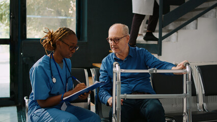 Female nurse doing checkup consultation with old patient in hospital waiting area, having walking frame. Medical assistant writing clinical report or appointment form, health center.