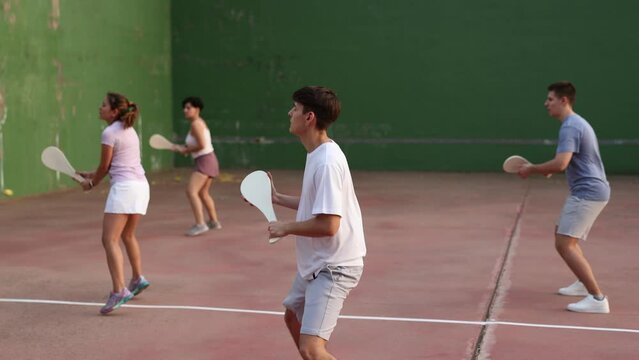 Group of sporty young people serving ball with special equipment, wooden paleta, during Basque pelota game outdoors. Boy playing pelota on outdoor fronton.