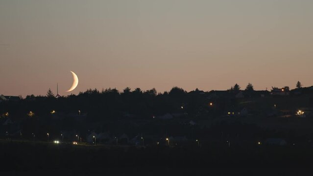 Sunset shot of a crescent, blood moon as it sets over Stornoway. Cars, a wind farm and houses are visible in the frame. Shot on the Isle of Lewis, part of the Outer Hebrides of Scotland.