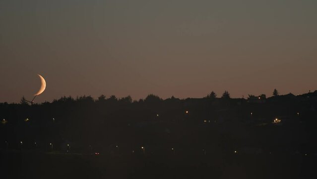 Evening shot of a crescent, blood moon setting over Stornoway. Cars, a wind farm and houses are visible in the frame. Shot on the Isle of Lewis, part of the Outer Hebrides of Scotland.