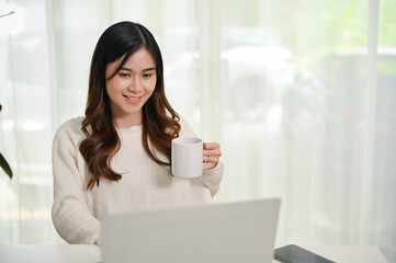 Attractive Asian woman having coffee and using her laptop at her home workspace.