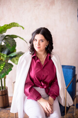 Fototapeta na wymiar A young girl stylist with dark hair smiles in a trendy white jacket, a burgundy color shirt and light trousers in a light studio with green living plants in pots 