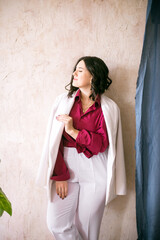 A young girl stylist with dark hair smiles in a trendy white jacket, a burgundy color shirt and light trousers in a light studio with green living plants in pots

