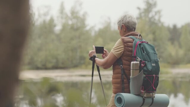 Medium shot of active senior Asian woman taking photos on smartphone of beautiful landscape while hiking by water in forest