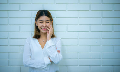 Beautiful asian business woman is standing with one hand on her face smiling while closing her eyes with white bricks in the background, Digital marketing.